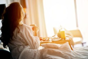 Kvinna i vit morgonrock äter frukost på sängen i ett ljust hotellrum.