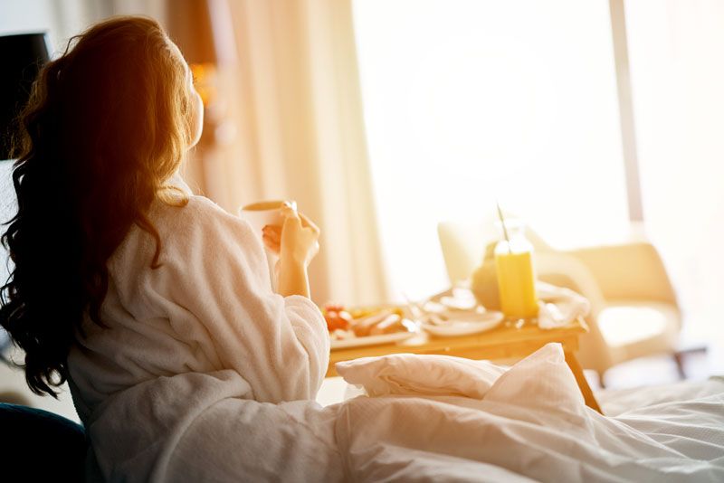Kvinna i vit morgonrock äter frukost på sängen i ett ljust hotellrum.